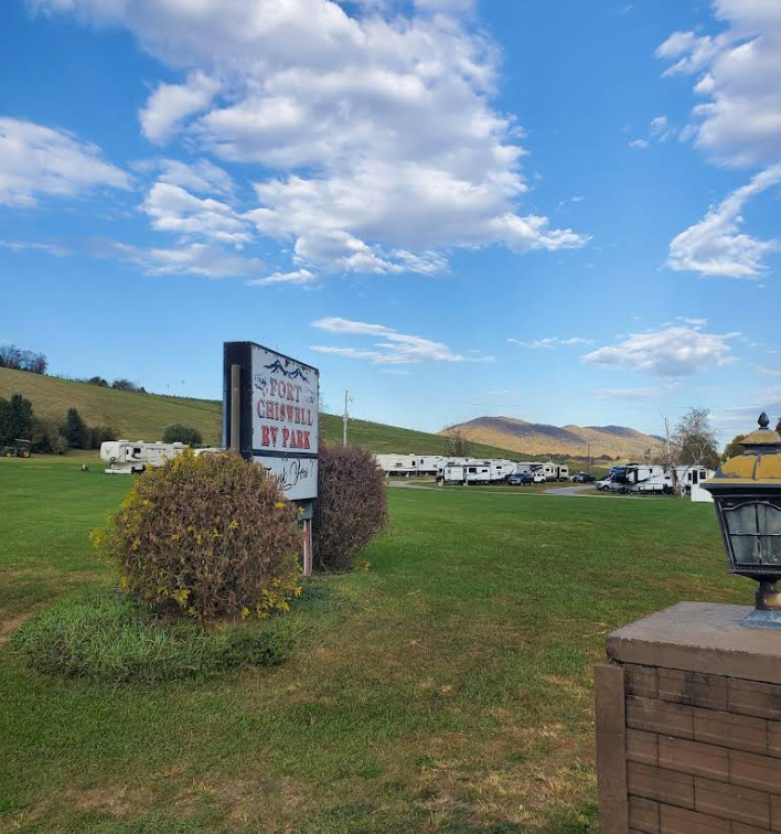 A grassy RV park with several trailers and a sign reading "Fort Chiswell RV Park" under a blue sky with scattered clouds.