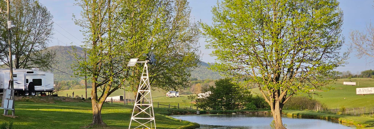 Two leafy trees stand beside a small pond with a white windmill and a camper nearby; grassy hills and distant mountains are visible in the background.